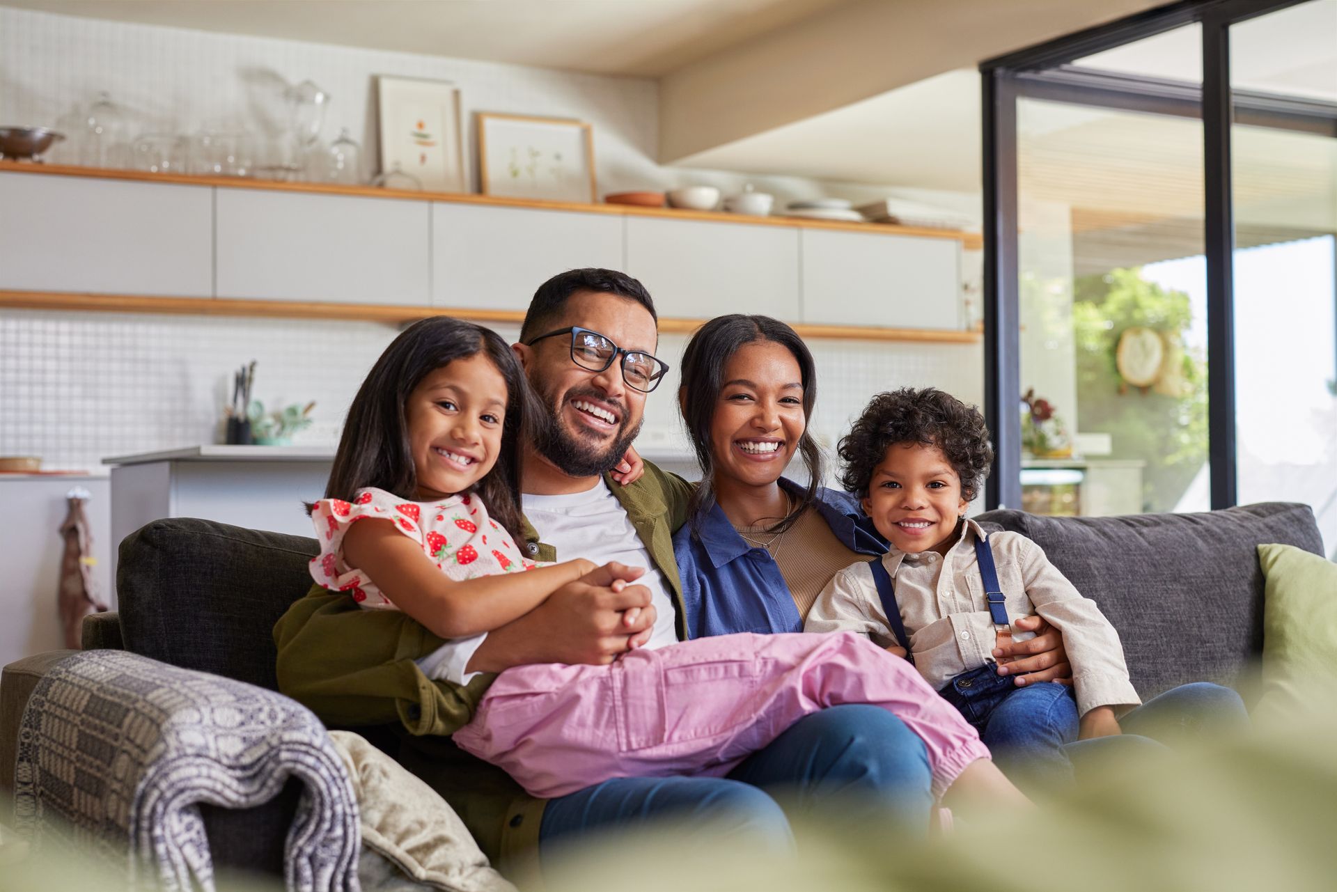 Family smiling on a couch in a bright living room, with a child in pink pants in front.