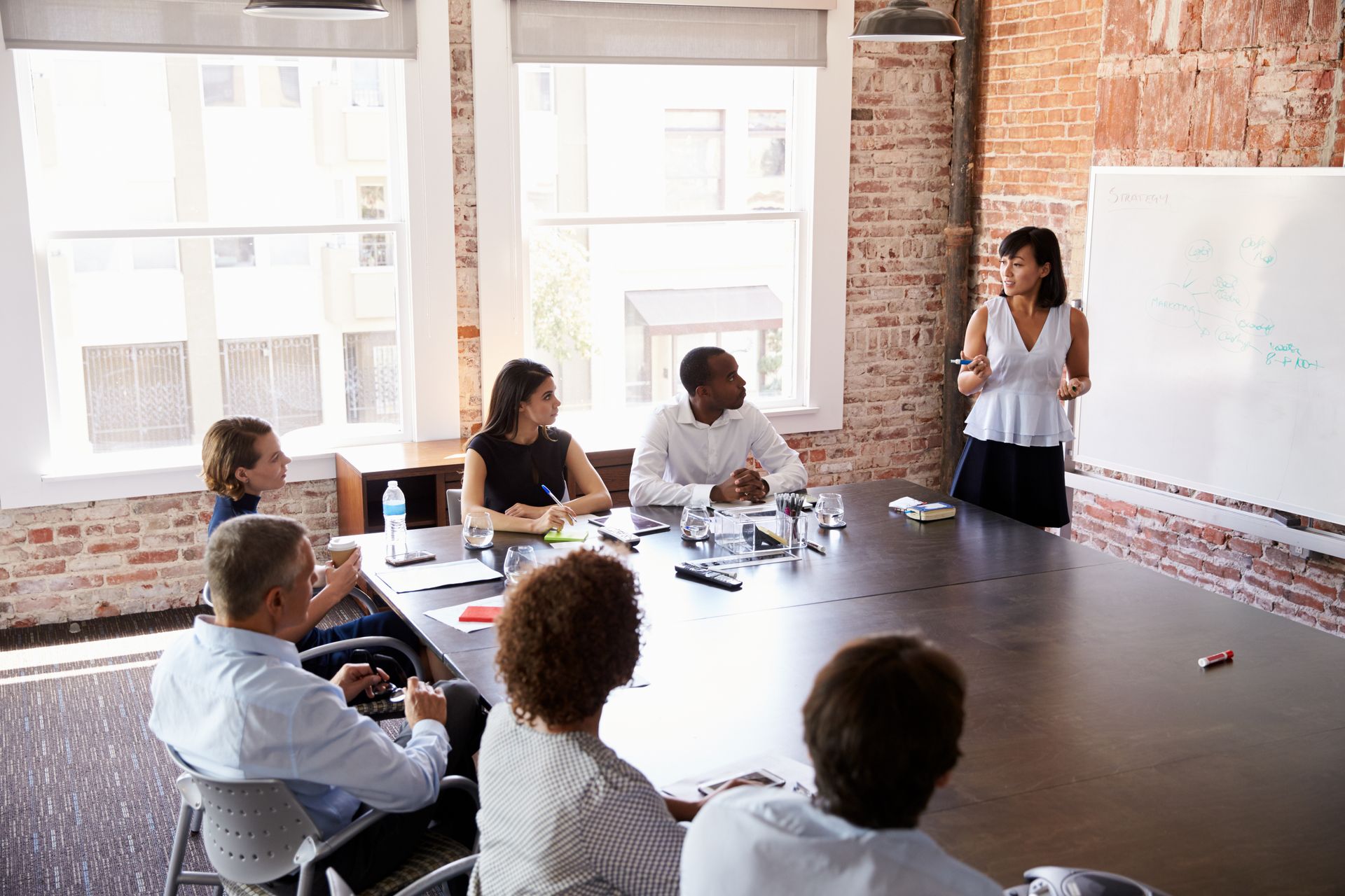 Meeting room with a presenter speaking to a small group around a conference table and whiteboard
