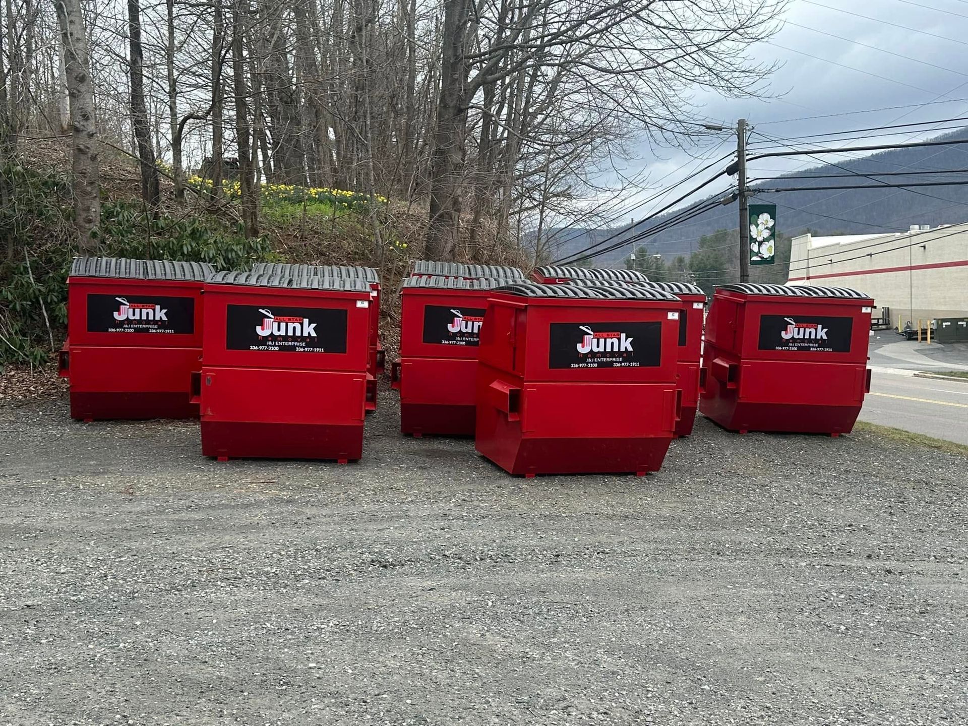Four red junk dumpsters lined up on gravel in front of bare trees and a building