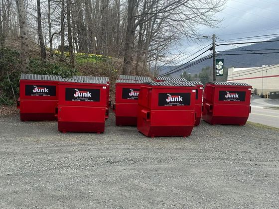Four red junk dumpsters lined up on gravel in front of bare trees and a building