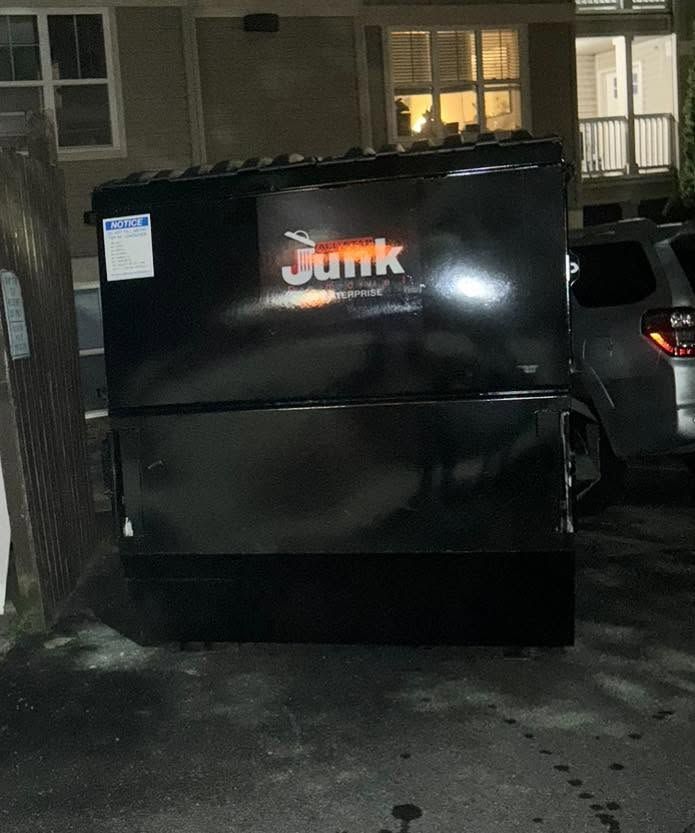 Black junk container in a driveway at night, with a lit house and parked car behind it.