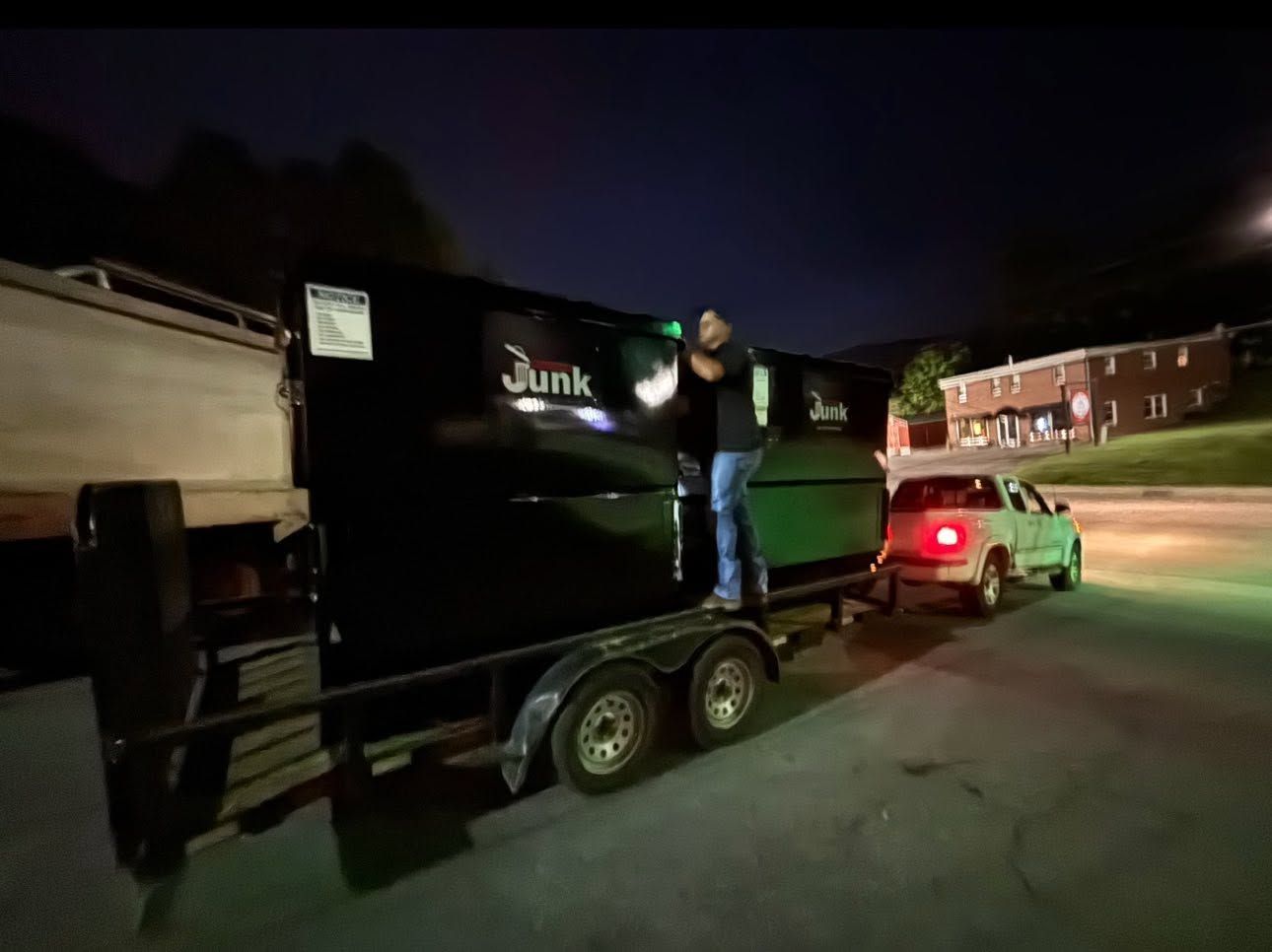 Person standing on a black junk trailer hitched to a car at night, with a red brick building in the background