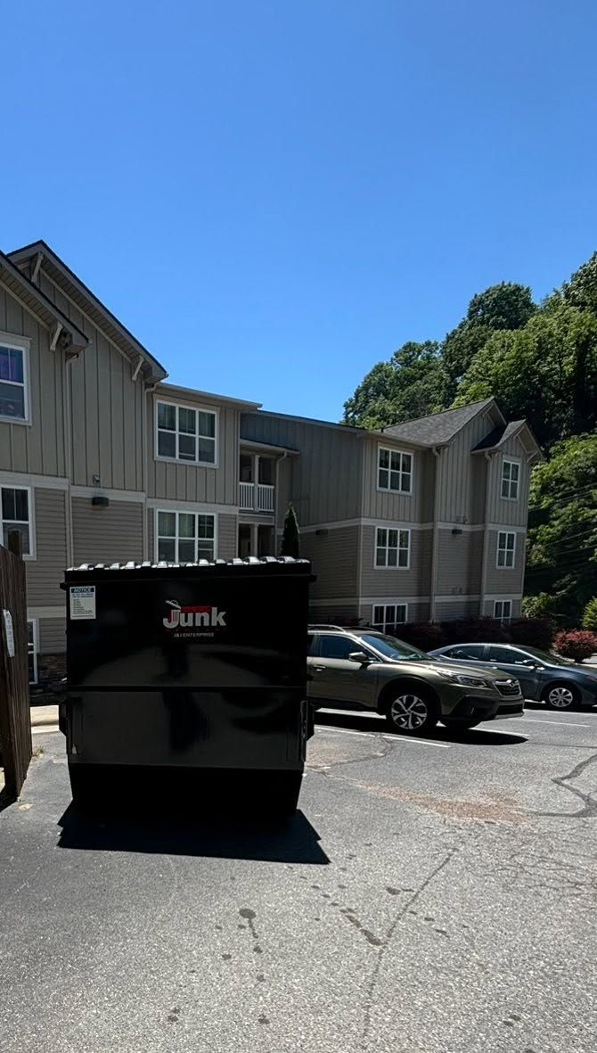 Apartment buildings, parked cars, and a black dumpster in a sunny parking lot under a clear blue sky