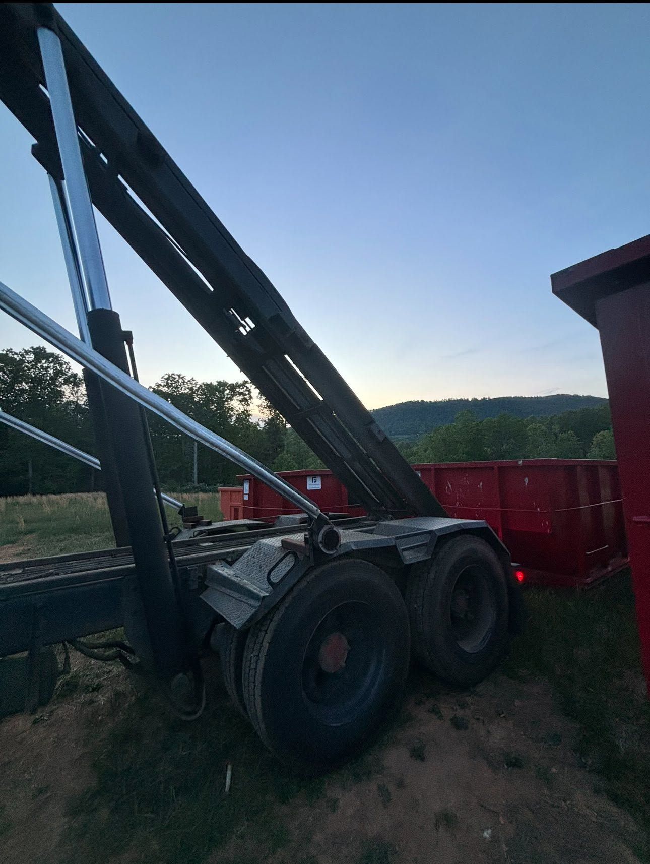 Crane truck with large black boom parked beside a red trailer in a grassy field at dusk