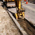 Excavator digging a trench beside a road, with dirt piled along the curb and traffic cones nearby.