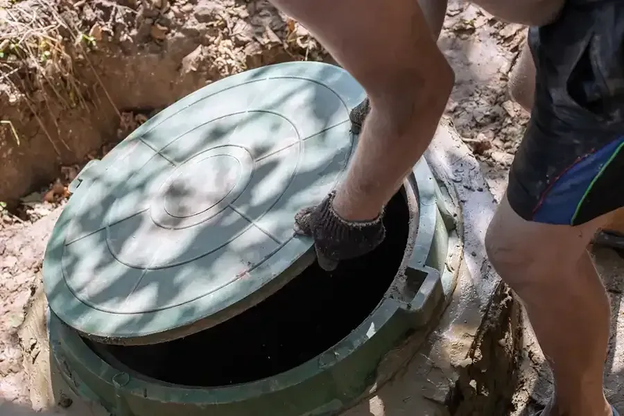 Person opening a round green manhole cover outdoors with gloves and shorts in a rocky area