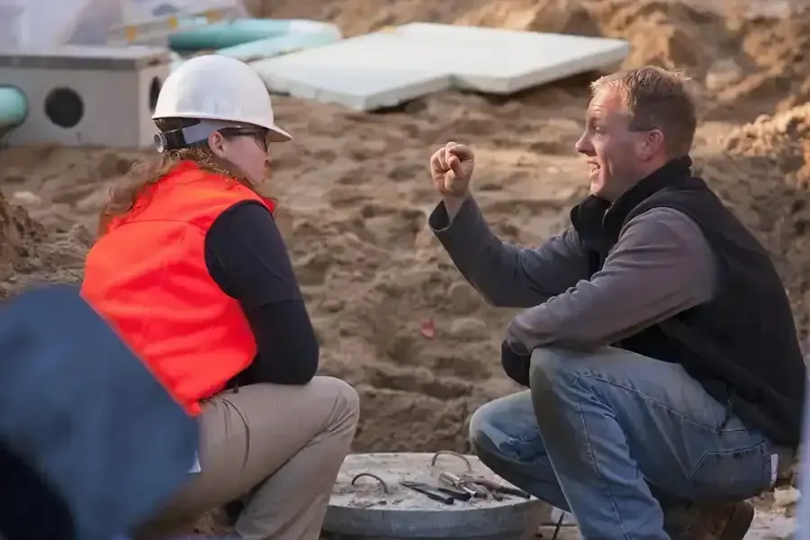 Two people in hard hats talking at a construction site, one holding up an object.