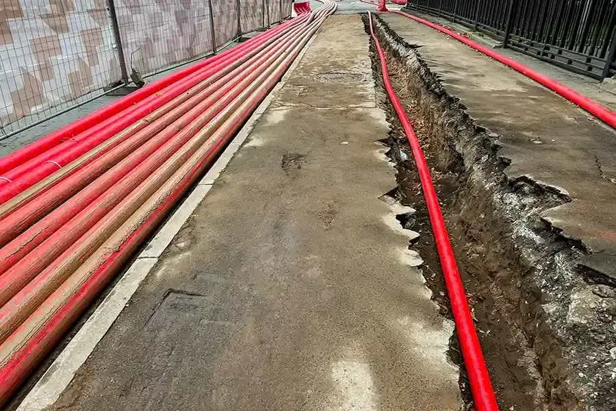 Red utility pipes laid along a long concrete trench beside a fence in a narrow outdoor corridor
