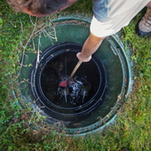 Person using a rod to inspect a black sewer manhole outdoors in grass