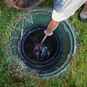 Person using a rod to inspect a black sewer manhole outdoors in grass
