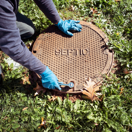 Gloved worker opening a round septic tank cover in grass with a wrench