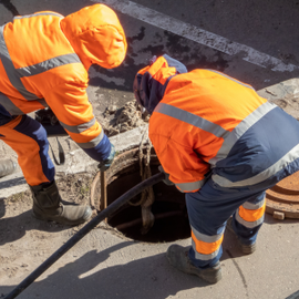 Two workers in orange safety gear inspect an open manhole on a street.