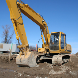 Yellow excavator with raised arm and bucket on a muddy construction site under a clear blue sky