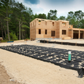 Partially built wooden house with foundation work and black ground cover in a wooded area