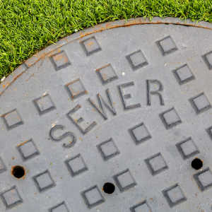 Round gray sewer manhole cover on grass, with raised square pattern and “SEWER” text.