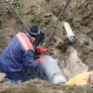 Utility worker repairing a buried pipe in a muddy trench with steam or dust rising nearby