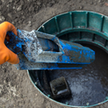 Orange-gloved hand holding a mud-covered blue metal tool over an open underground utility hole
