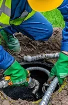Gloved worker inserting a gray hose into a ground drainage opening on grass