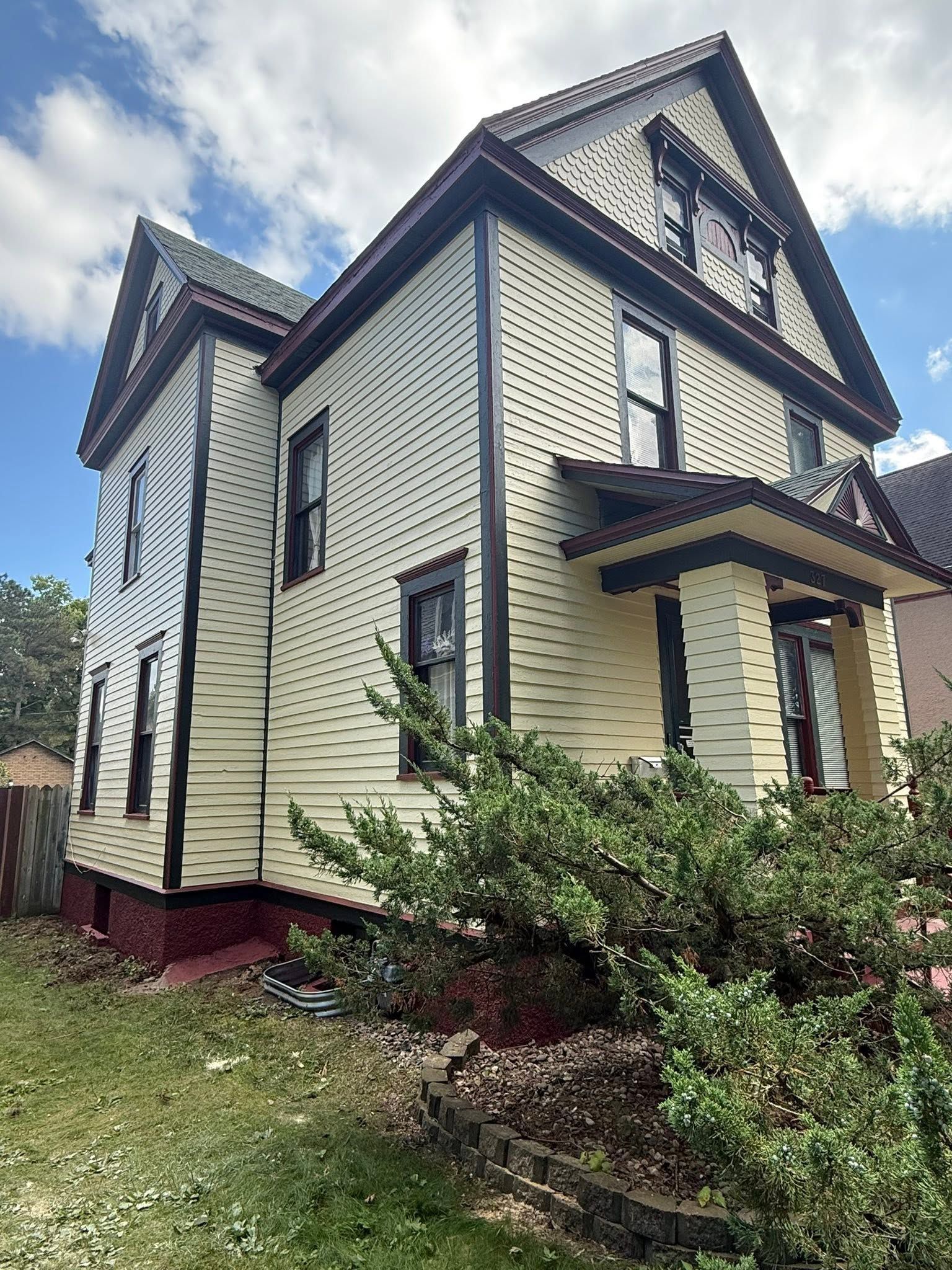 Victorian-style cream house with dark trim, viewed from the front amid shrubs and grass