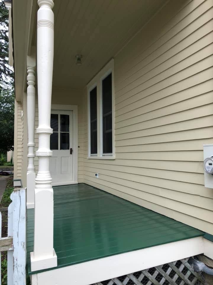 Front porch with green floor, white trim, beige siding, and a white door with windows