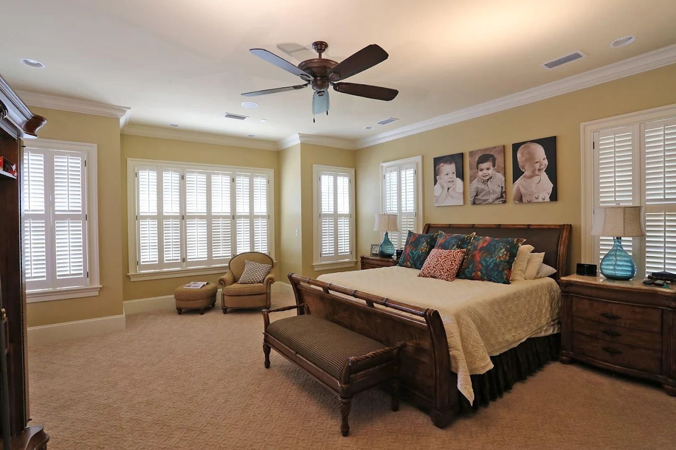 Spacious beige bedroom with a king bed, bench, armchairs, shuttered windows, and ceiling fan.