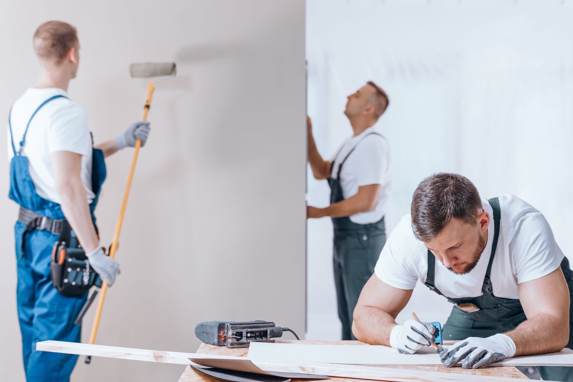 Three painters in white shirts and blue overalls working on a wall in a bright room