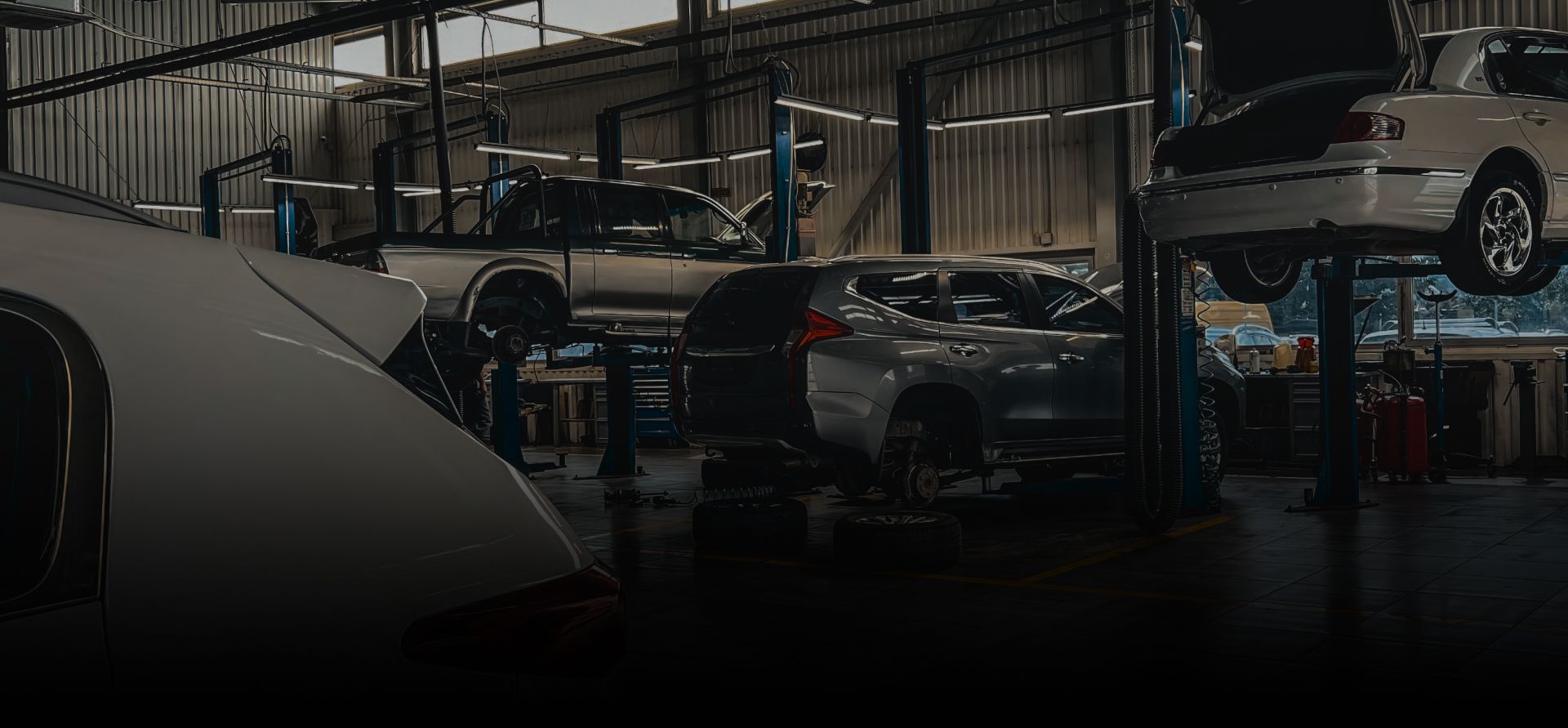 Luxury cars in a dim workshop, with a vehicle lifted on a repair rack and others parked below | Rick's Auto Shop