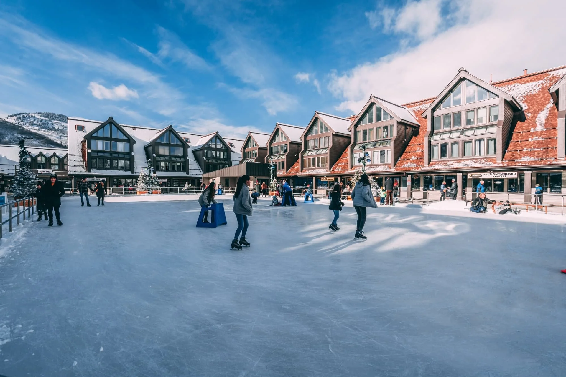 Ice skaters on an outdoor rink in front of buildings with red-tiled roofs on a sunny day.