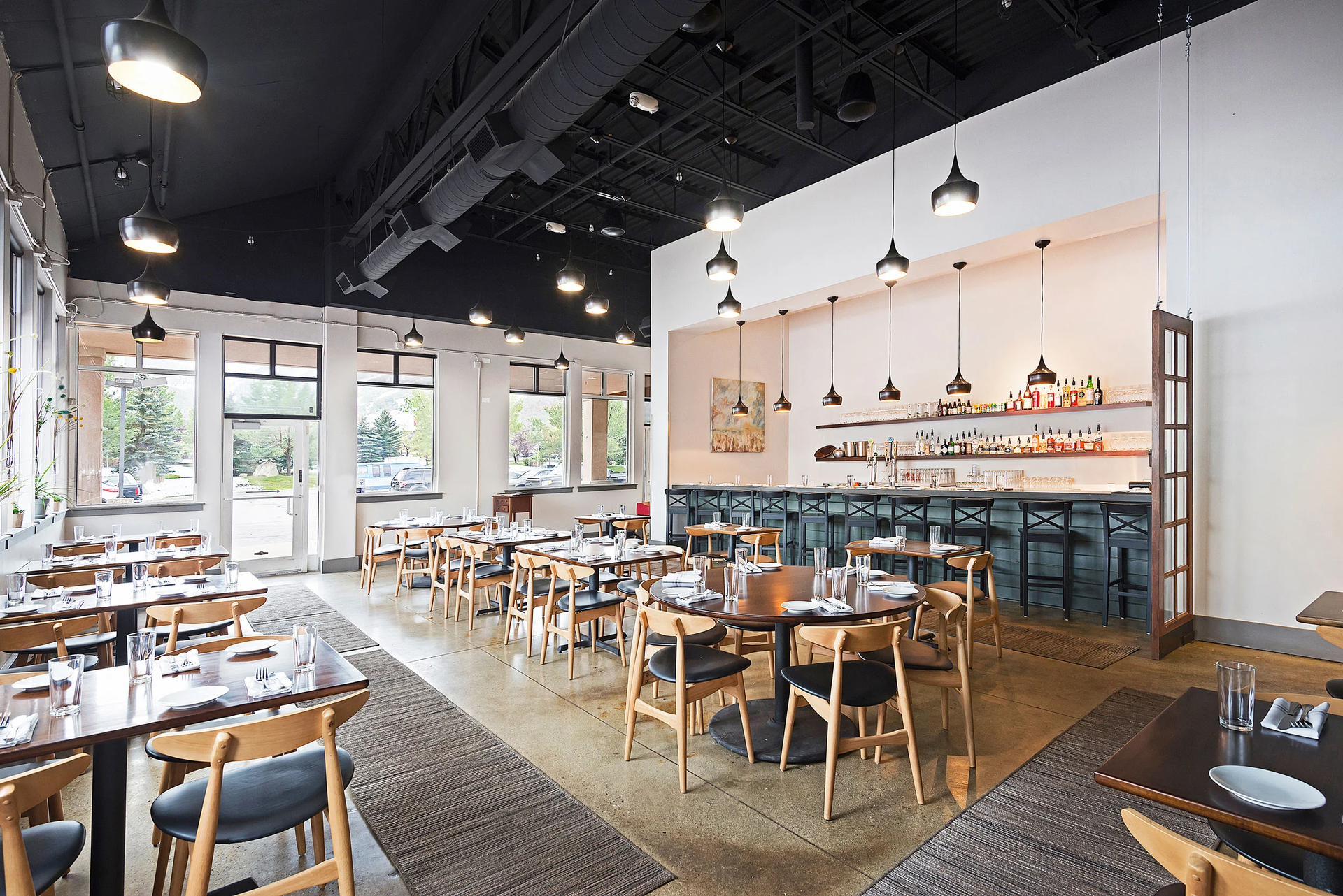 Interior of a modern restaurant with tables, chairs, bar, and hanging lights. White walls, dark ceiling.