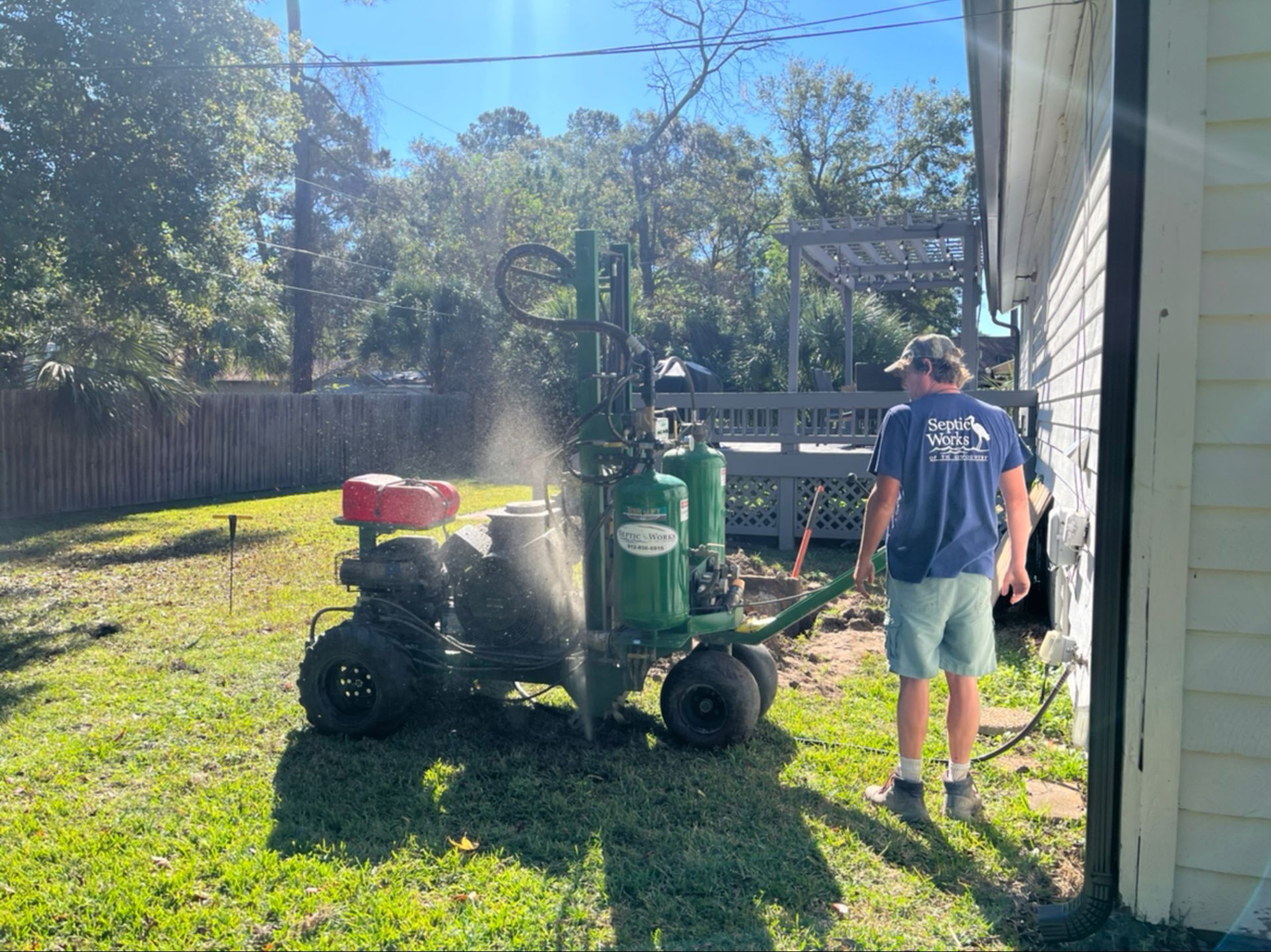 Man operating a green drilling machine next to a house; water spraying.