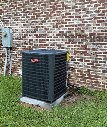 Black outdoor HVAC unit beside a brick wall and electrical box, on a concrete pad in a grassy yard