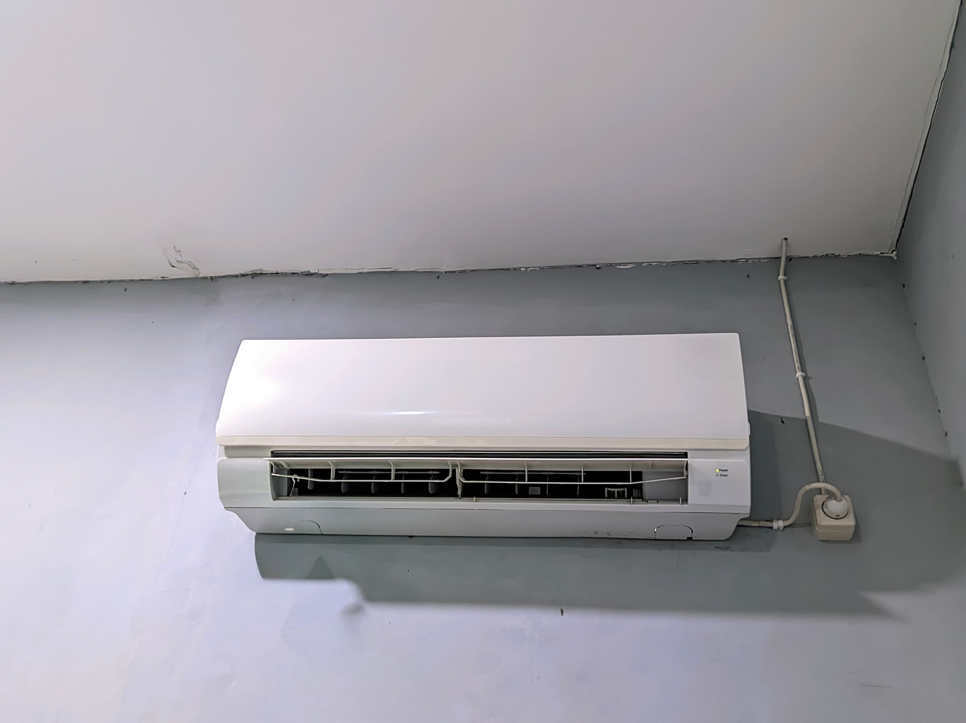 Wall-mounted white air conditioner near the ceiling with visible power cord on a gray wall