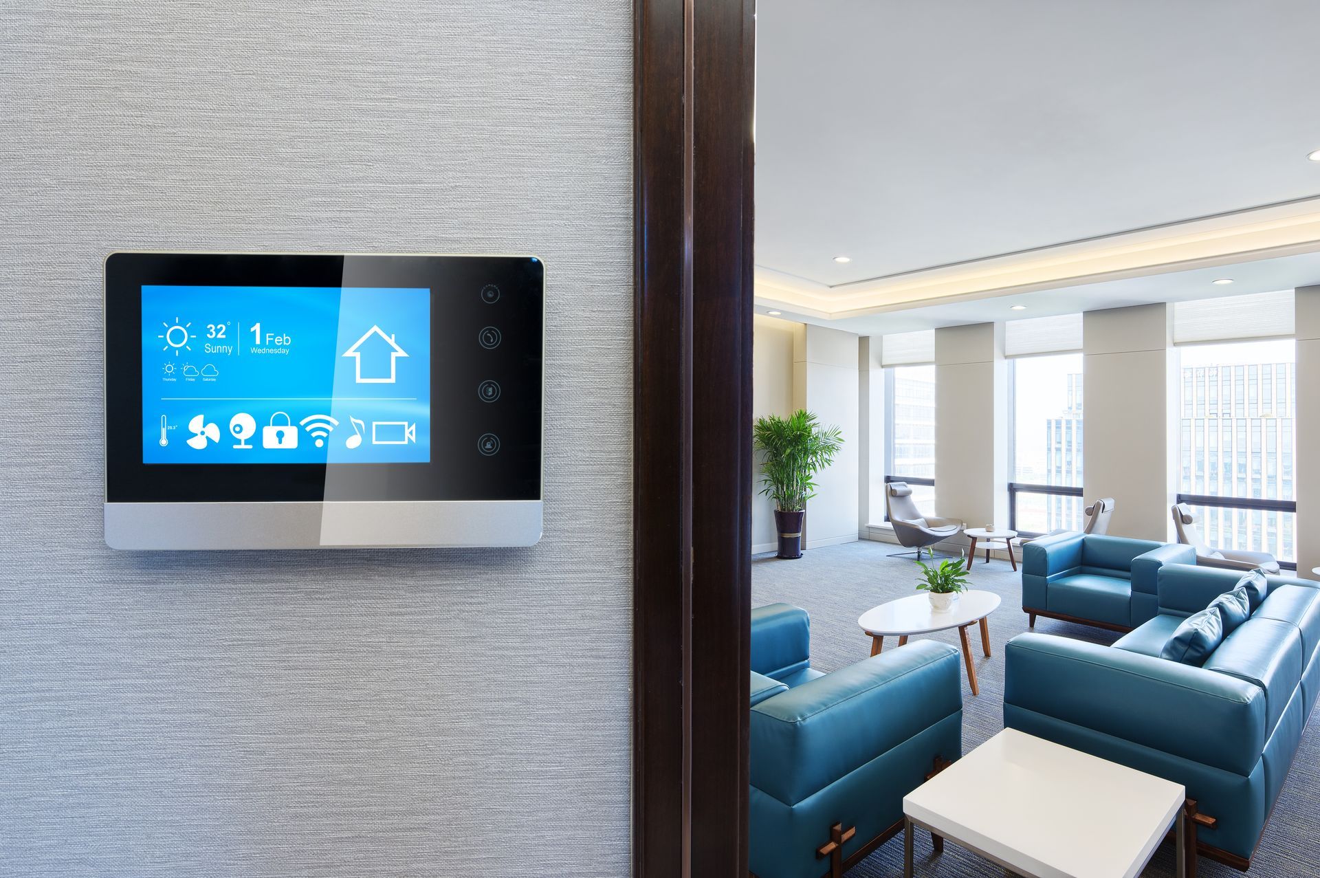 Wall-mounted touchscreen control panel beside a bright modern living room with blue chairs and sofa
