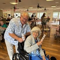 Man pushing a wheelchair with a smiling woman holding cards in a bright community room