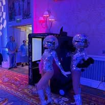 Two mirrored disco-costumed performers in a purple-lit hallway at a party