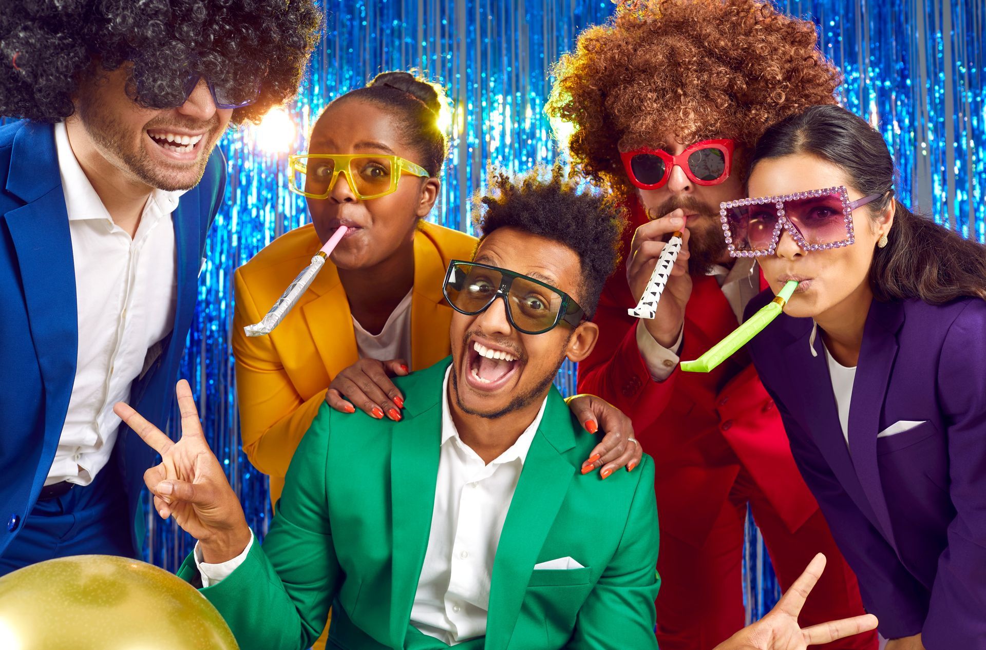 Five people in colorful suits and wigs pose for a party photo against a metallic blue background, smiling