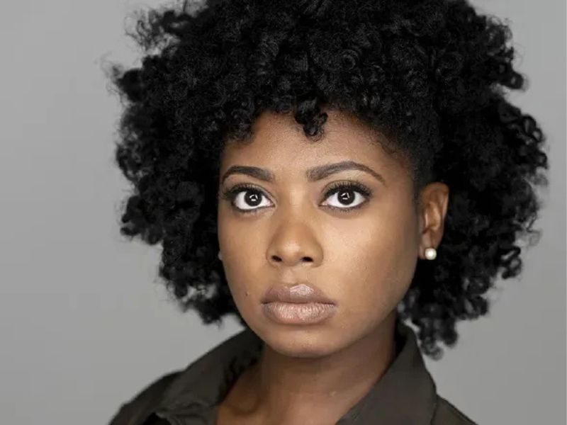 Portrait of a woman with curly black hair, neutral expression, and a dark jacket against a gray background