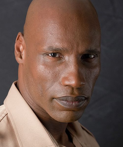 Close-up portrait of a bald man in a beige shirt against a dark background.
