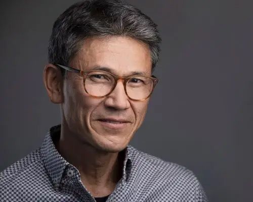 Portrait of a man with glasses and gray hair, wearing a patterned shirt against a dark background
