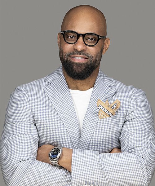 Portrait of a bald man with glasses, beard, light plaid blazer, white shirt, and folded arms on a gray background