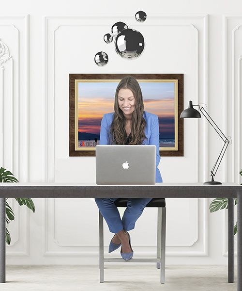 Person working on a laptop at a desk in a bright office with a framed sunset painting behind them.