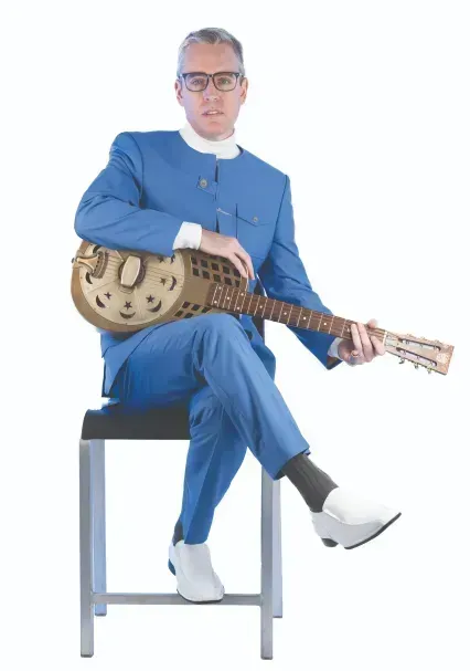 Man in a blue suit seated on a stool, holding a guitar against a white background
