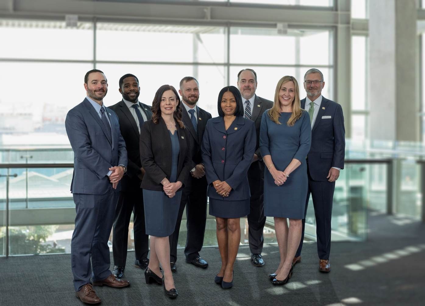 Seven professionals posing in business attire in a bright office lobby