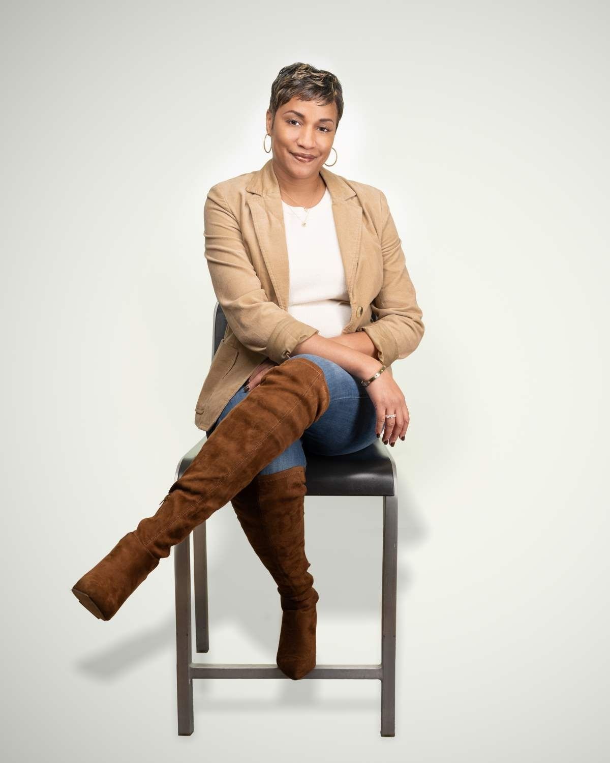 Person seated on a stool, wearing a tan blazer and tall brown boots against a light background.