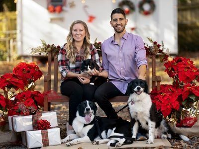 Two people seated with three dogs among red holiday decorations and wrapped gifts