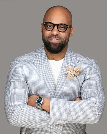 Portrait of a man in glasses and a gray checkered blazer, arms crossed against a gray background