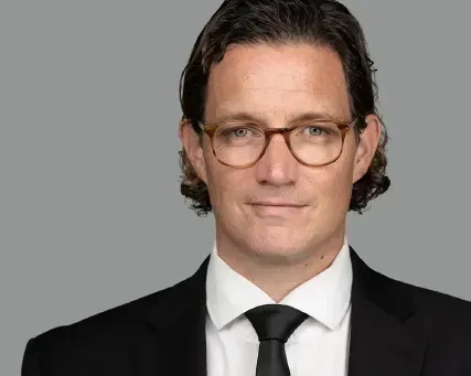 Head-and-shoulders portrait of a man in a black suit, white shirt, and tie against a gray background