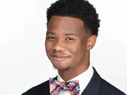 Smiling man in a dark suit and red plaid bow tie against a white background