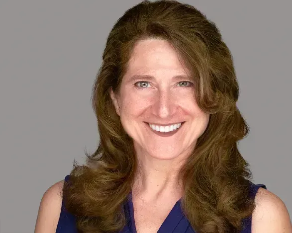 Smiling woman with wavy brown hair wearing a dark blue top against a gray background