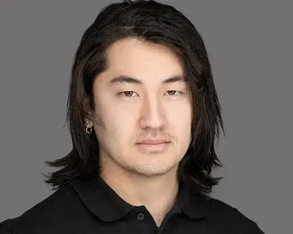 Portrait of a person with shoulder-length black hair, wearing a black shirt, against a gray background